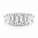 A three-stone diamond engagement ring featuring an emerald-cut center stone and baguette-cut side stones in a platinum metal setting, shown on a clean white background with a soft reflection.