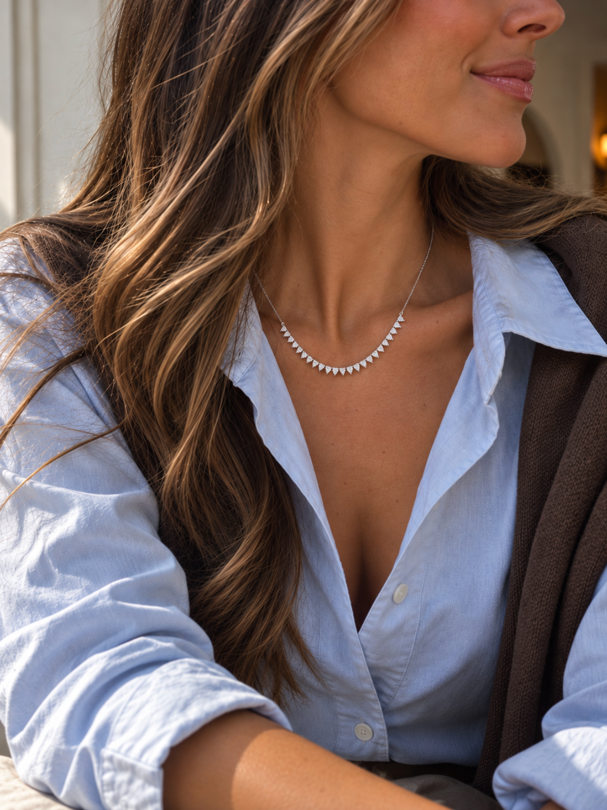 A close-up lifestyle shot of a woman wearing a delicate white gold necklace with evenly spaced diamonds. She is wearing a light blue button-down shirt and a brown cardigan, with her hair styled in soft waves.
