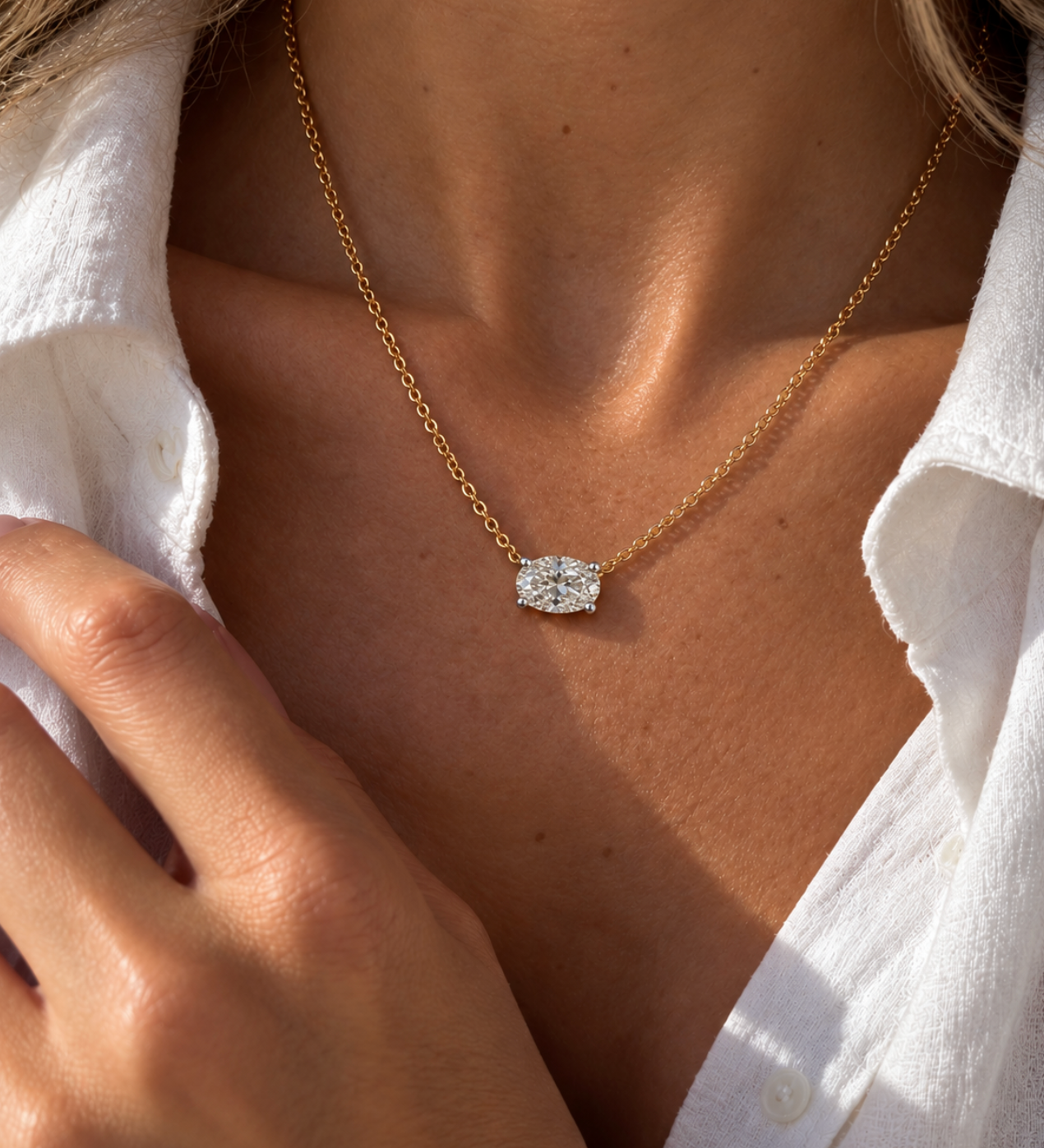 A close-up lifestyle shot of a woman wearing a delicate gold chain necklace with a oval diamond pendant, styled with a white button-down shirt.