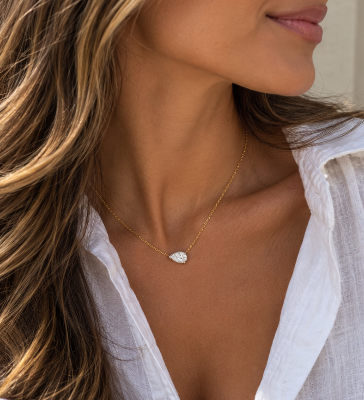 A close-up lifestyle shot of a woman wearing a delicate gold chain necklace with a pear cut diamond pendant, styled with a white button-down shirt.