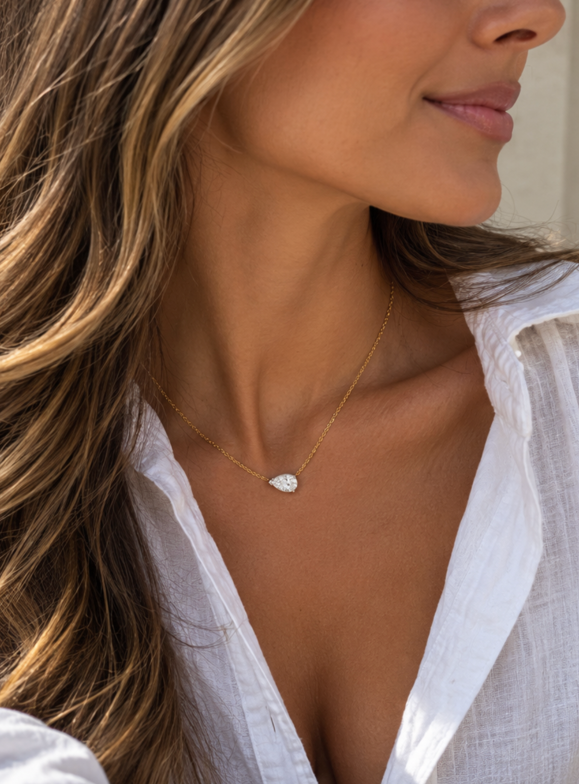 A close-up lifestyle shot of a woman wearing a delicate gold chain necklace with a pear cut diamond pendant, styled with a white button-down shirt.