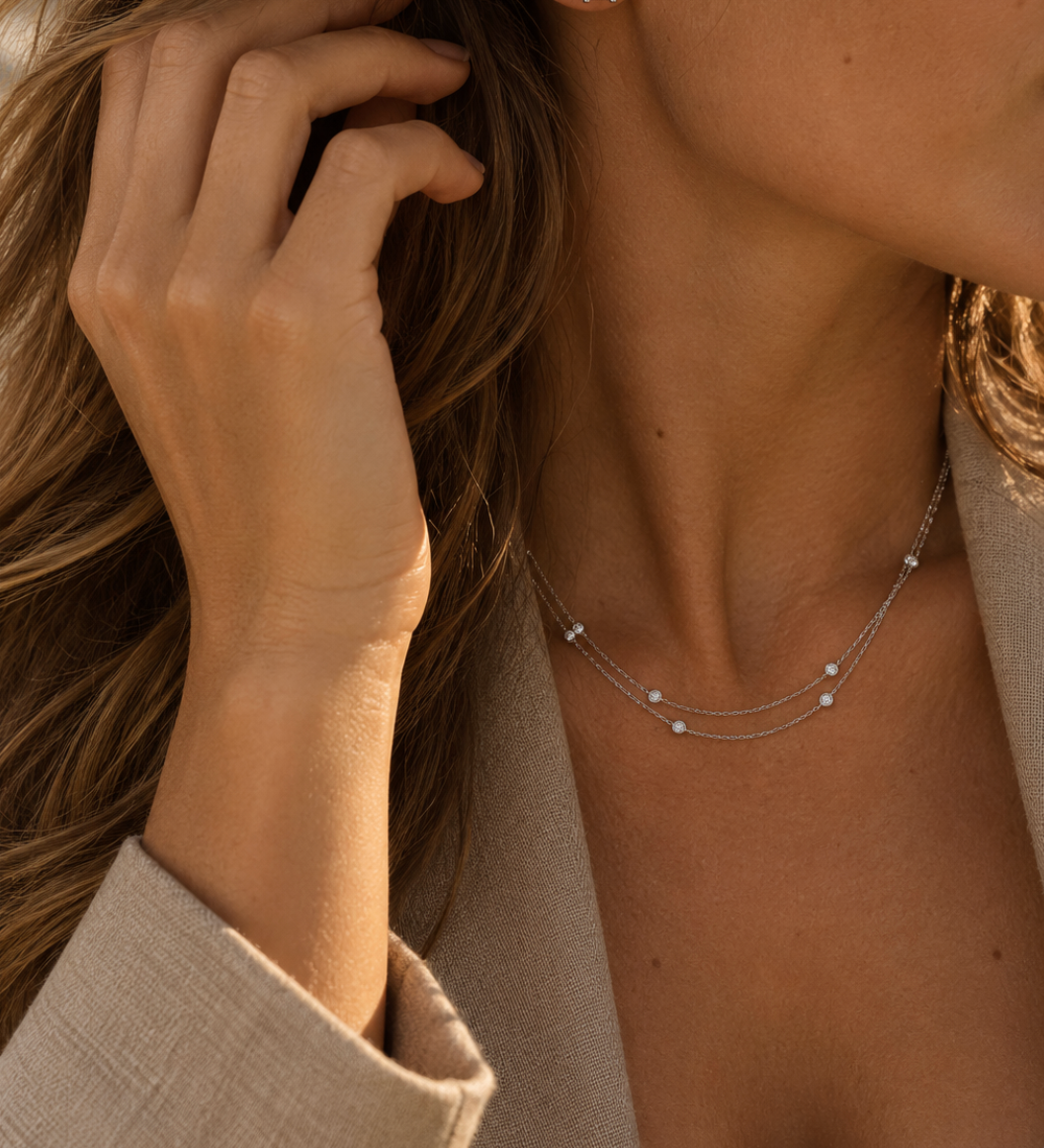 A close-up lifestyle shot of a woman wearing a delicate double-strand white gold necklace with small diamonds and a matching square stud earring with a diamond gemstone.