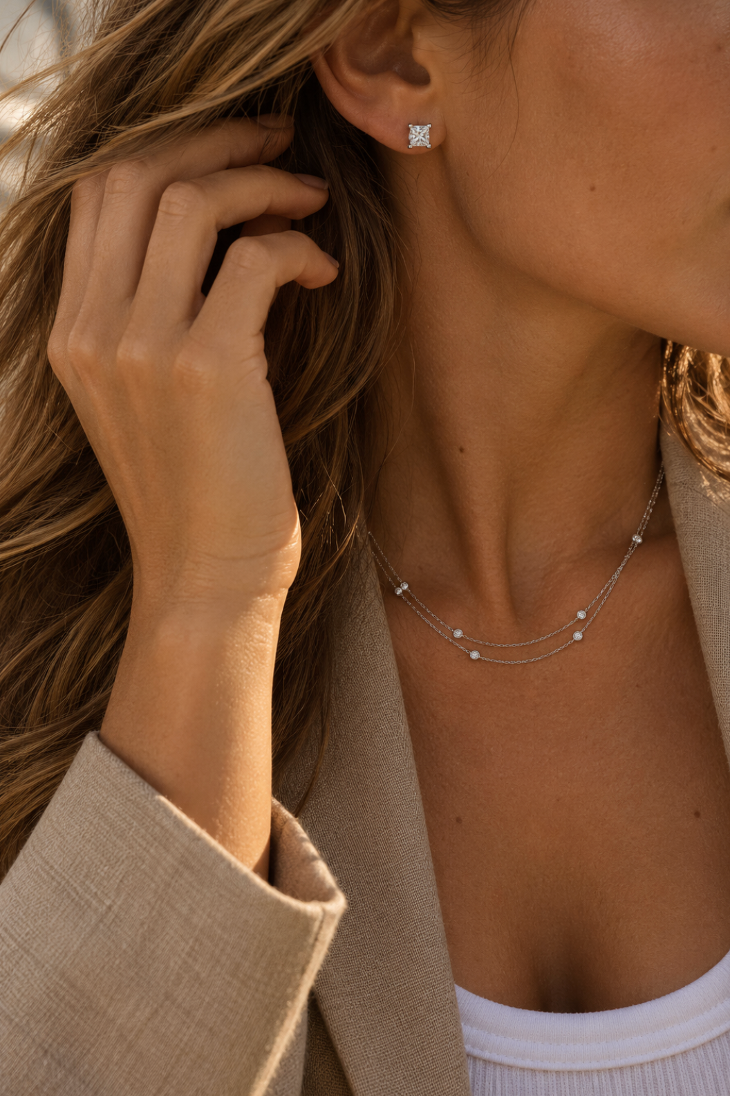 A close-up lifestyle shot of a woman wearing a delicate double-strand white gold necklace with small diamonds and a matching square stud earring with a diamond gemstone.