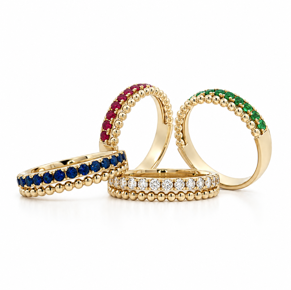 Four gold eternity rings arranged on a white background, each featuring a different colored gemstone band: red, blue, green, and clear stones.