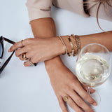 Close-up of a woman's hands with jewelry holding a glass of white wine on a light surface.