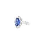 18ct white gold ring featuring a 5.11ct emerald cut tanzanite framed by emerald and round brilliant cut diamonds in a detailed halo setting with upswept shoulders.
