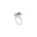 18ct white gold ring featuring a 5.11ct emerald cut tanzanite framed by emerald and round brilliant cut diamonds in a detailed halo setting with upswept shoulders.