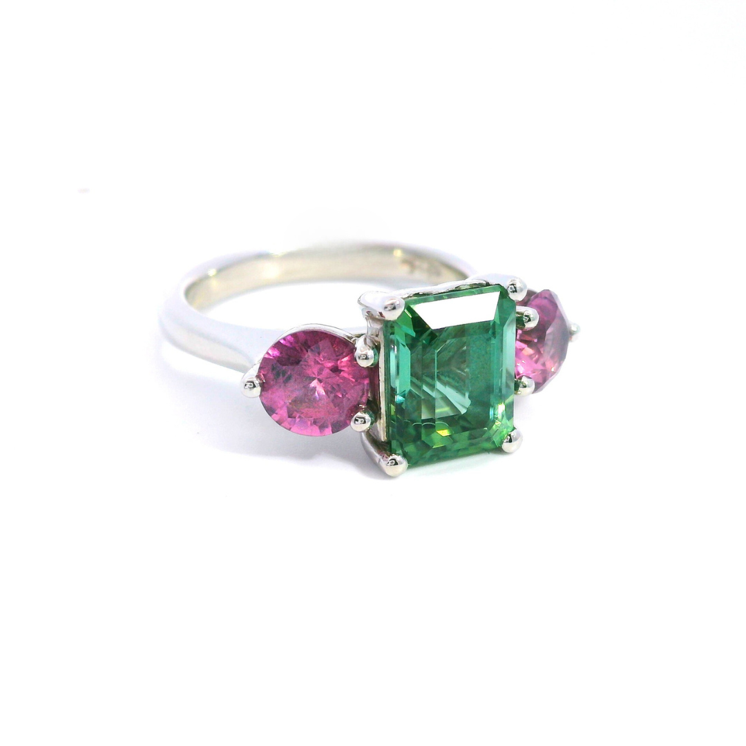 Platinum three-stone ring featuring a 2.68ct emerald cut green tourmaline with two round cut pink spinels in tiered claw settings.