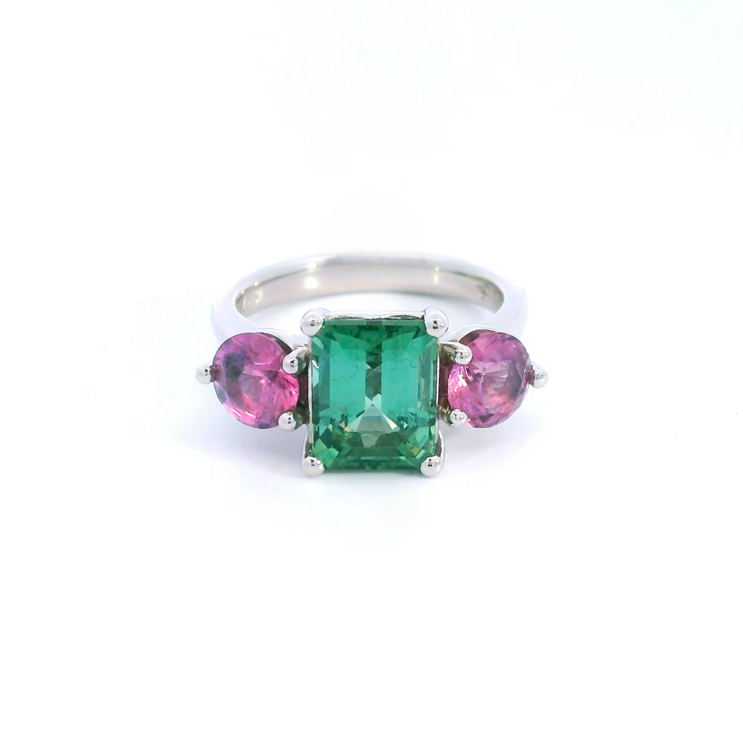Platinum three-stone ring featuring a 2.68ct emerald cut green tourmaline with two round cut pink spinels in tiered claw settings.