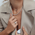 Person wearing a beige trench coat with a necklace and ring, holding the coat open. 18ct white gold three stone diamond ring featuring a 2.17ct oval centre diamond with pear cut diamond side stones in tiered cap-and-claw settings.