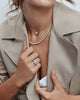 Person wearing a beige trench coat with a necklace and ring, holding the coat open. 18ct white gold three stone diamond ring featuring a 2.17ct oval centre diamond with pear cut diamond side stones in tiered cap-and-claw settings.