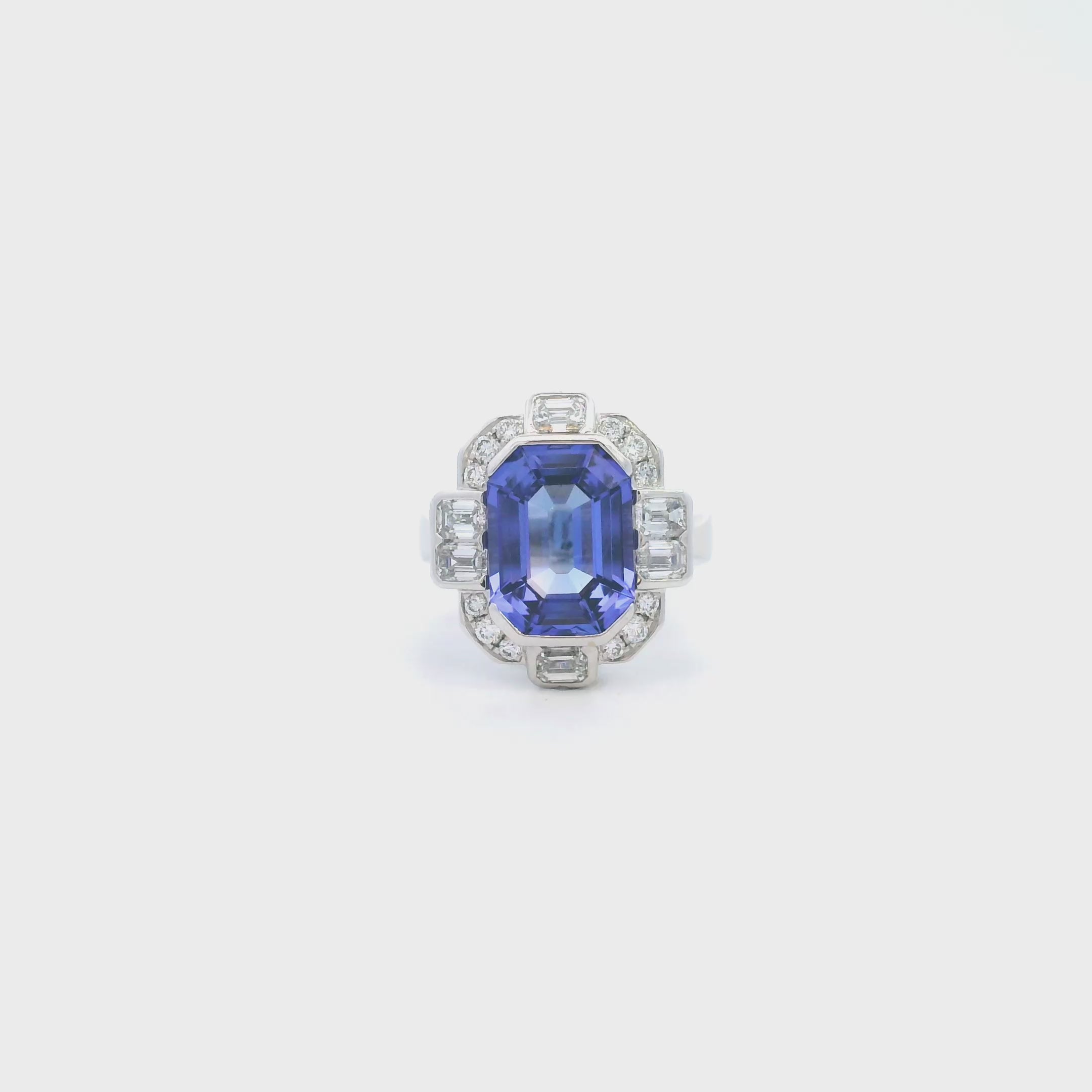 18ct white gold ring featuring a 5.11ct emerald cut tanzanite framed by emerald and round brilliant cut diamonds in a detailed halo setting with upswept shoulders.