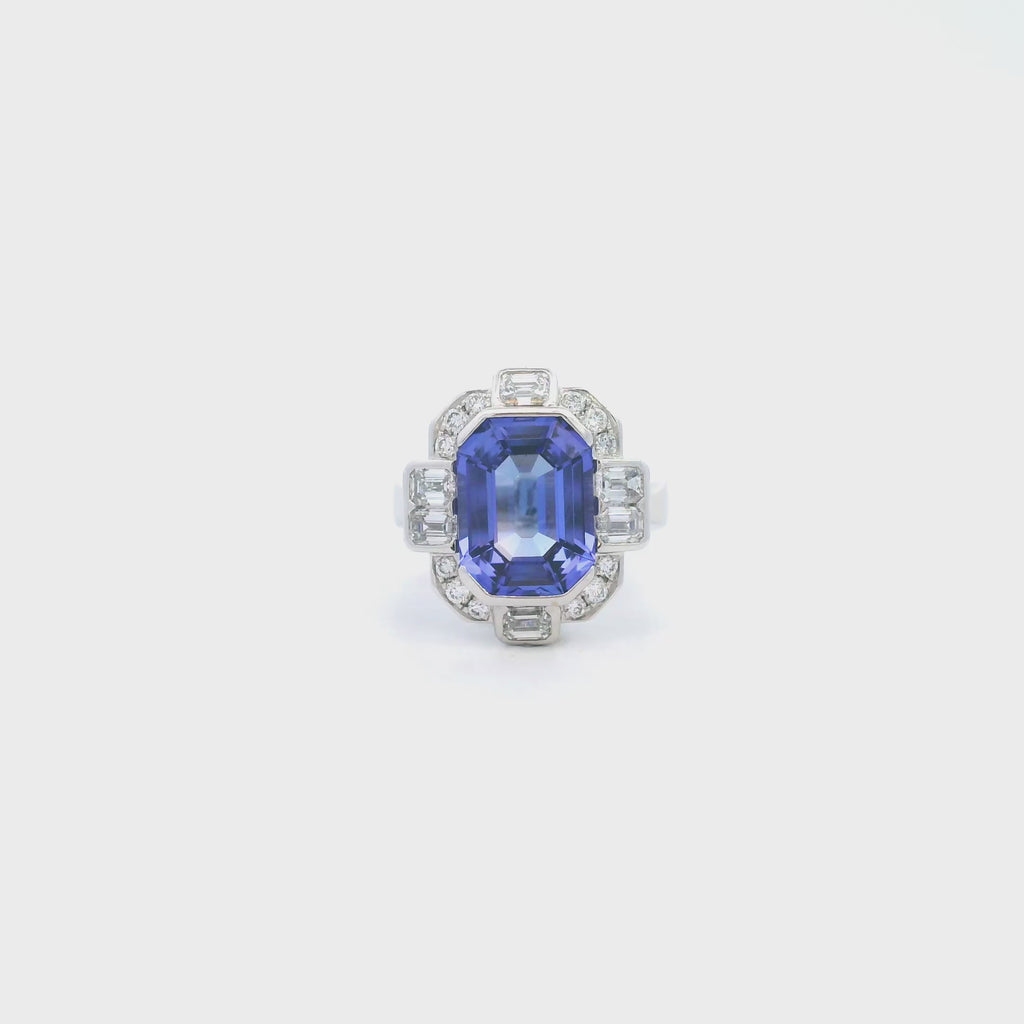 18ct white gold ring featuring a 5.11ct emerald cut tanzanite framed by emerald and round brilliant cut diamonds in a detailed halo setting with upswept shoulders.